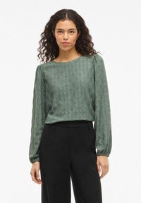 Zelená vzorovaná blůza s dlouhými rukávy, kulatým výstřihem a strukturovanou tkaninou. V kombinaci s černými upravenými kalhotami modelka stojí na bílém pozadí.