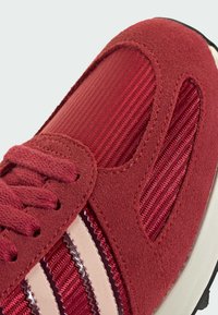 Κόκκινο αθλητικό παπούτσι με συνδυασμό μαλακού σουέντ και υφασμάτινης υφής, με τρεις κρεμ ρίγες και κλείσιμο με κορδόνια στο  επάνω μέρος.