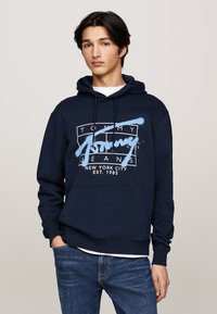 Sudadera azul marina de algodón con un bolsillo frontal y capucha ajustable. Presenta un logo gráfico blanco que dice "Tommy Jeans" y "Nueva York".