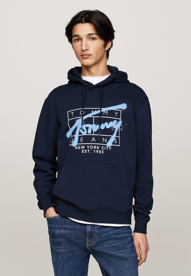 Sudadera azul marina de algodón con un bolsillo frontal y capucha ajustable. Presenta un logo gráfico blanco que dice "Tommy Jeans" y "Nueva York".