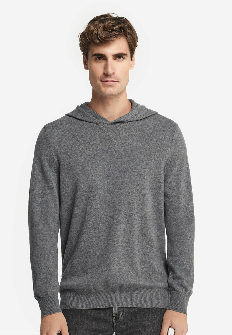 Junger Mann mit hellbraunen Haaren, der einen schlichten grauen Kapuzenpullover aus Strick und dunkelgraue Hosen trägt, stehend vor weißem Hintergrund.