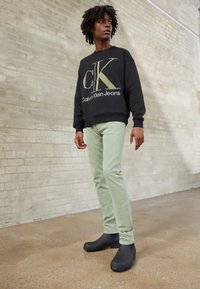 Black sweatshirt with large gold Calvin Klein logo, mint green jeans, and black rubber clogs on a textured floor against a beige wall.