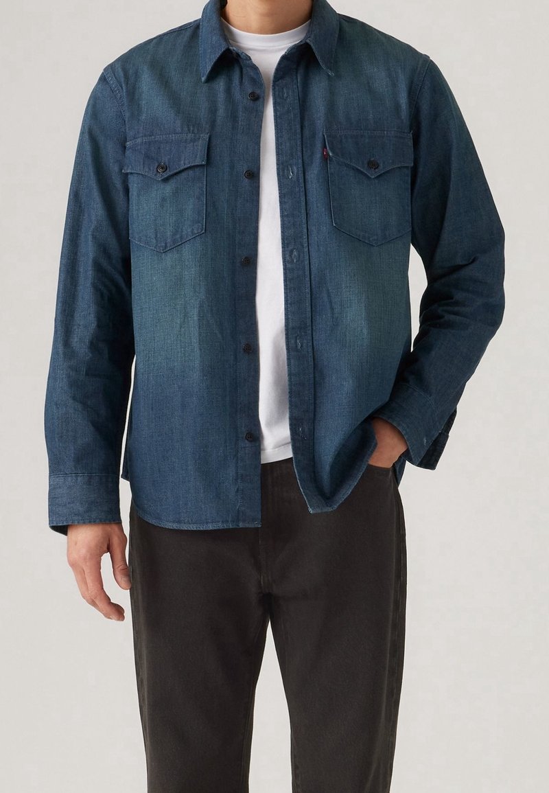 Homme portant une chemise en jean bleu foncé boutonnée par-dessus un t-shirt blanc avec un pantalon foncé, une main dans la poche, sur un fond uni.