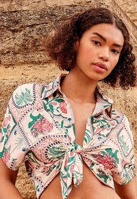 Jeune femme aux cheveux courts et bouclés portant une chemise à motifs floraux nouée, debout devant un mur en pierre texturé.