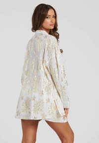 Prenda blanca con patrones de coral dorado y estrellas de mar. Mangas largas, corte holgado y abertura en la espalda. Tejido ligero y texturizado.