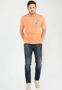 Camiseta naranja de manga corta con un bolsillo texturizado gris, combinada con vaqueros azul oscuro y zapatillas de color claro. El modelo está de pie con los brazos relajados.