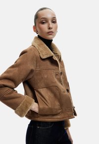 Brun imitation ruskindsjakke med en shearlingkrave og -manchetter, med knaplukninger og sidelommer, båret over en sort turtleneck.