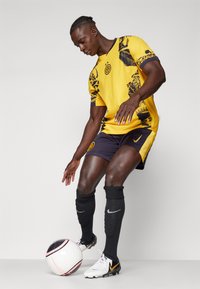 Kollane jalgpallisärk mustade aktsentide ja mustritega, tumesinised lühikesed püksid, mustad sukad ning valged saapad kollase logoga, hoiab jalgpalli.