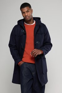 Cappotto di lana blu navy con cappuccio, dotato di chiusure a scorrimento e tasche laterali, indossato sopra un maglione arancione, con pantaloni di jeans scuri.
