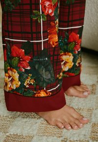 Floral printed pajama pants in red and green, with a plaid pattern, featuring a solid burgundy cuff at the hem. Bare feet visible.