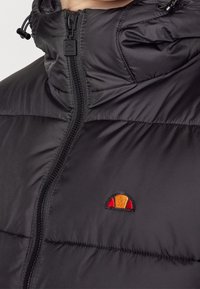 Musta topattu puhvutakki korkealla kauluksella, etuvetoketjulla ja pienellä puolikuun muotoisella logolla oranssina ja punaisena rinnassa.