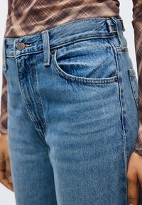 Άτομο που φορά μπλε τζιν και καφέ πουκάμισο με καρό σχέδιο μακρυμάνικο, που δείχνει την περιοχή της μέσης και τους κορυφαίους μηρούς.