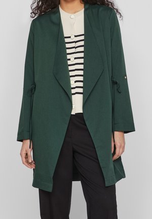 Kvinde iført mørkegrøn åben-frakke over hvid og sort stribet skjorte med knapper, kombineret med sorte bukser, stående foran en grå baggrund.