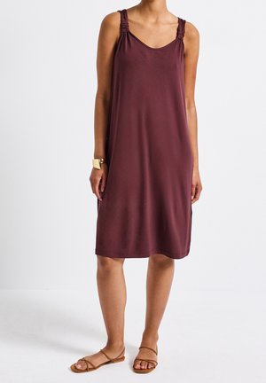 Kvinde iført en ærmeløs, knælængde vinrød kjole med rynkede stropper, brune sandaler med stropper og et guldfarvet armbånd på venstre håndled.