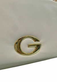 Superficie in pelle crema con un grande emblema dorato a forma di lettera G al centro, con una tracolla a catena parzialmente visibile nell'angolo superiore.