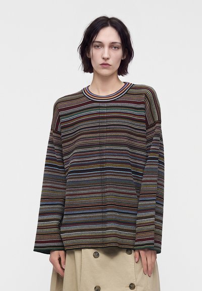 Jeune personne aux cheveux courts et foncés portant un pull rayé multicolore et une jupe beige boutonnée devant un fond clair uni.