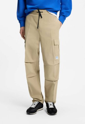Pantalon cargo beige avec poches latérales et cordon noir, porté avec des baskets noires et blanches et un sweat bleu.