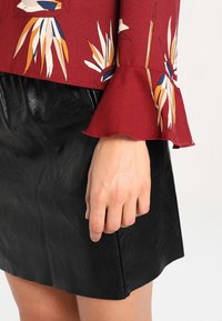 Bordeaux bluse med blomstermønster og flæseærmer, parret med en sort faux læder mini nederdel. Glat tekstur og tætsiddende design.