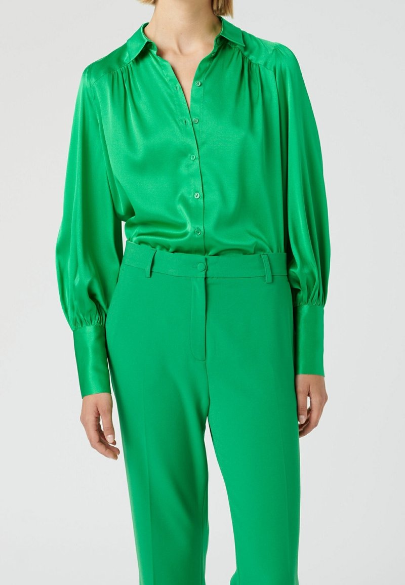Blusa verde in satin con colletto, apertura con bottoni e dettagli sulle spalle arricciati, abbinata a pantaloni verde a vita alta coordinati.
