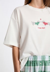 Biała koszulka z dwoma szarymi dachshundami w swetrach świątecznych i napisem "Przytulne dni", wsunięta w zielono-białe kratkowane spodnie.