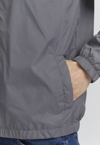 Grå letvægtsjakke lavet af glat nylon, med en sidelomme, elastiske manchetter og en kant der sidder tætsiddende omkring kroppen.