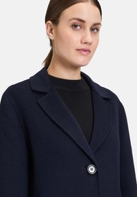 Marineblå uldblandingsfrakke med reverskrave, enkelt knaplukning og glat tekstur. Bæres over en sort sweater for lagdeling.