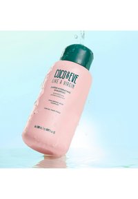 Rosa schampuflaska med en mörkgrön kork, märkt "Coco & Eve Like a Virgin Super Hydrating Shampoo," med vattendroppar på ytan.