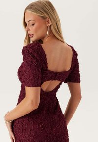 Burgundy klänning med blommönster, korta ärmar och skärning i ryggen. Slät textur med en figurnära silhuett och dold dragkedja.