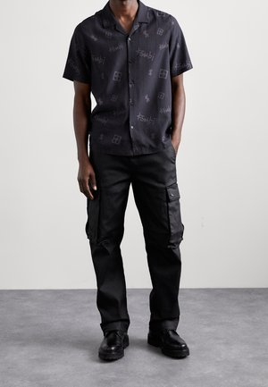 Camicia nera a maniche corte con bottoni e un motivo sottile, abbinata a pantaloni cargo neri con tasche laterali e calzature robuste.