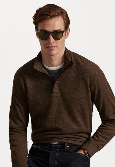 Καφέ πουλόβερ με φερμουάρ και σχέδιο ψαροκόκκαλου, φτιαγμένο από υφασμάτινη υφή, τοποθετημένο πάνω από γκρι πουκάμισο, συνδυασμένο με σκούρα τζιν και γυαλιά ηλίου.