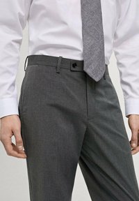 Pantaloni gri din țesătură netedă, cu față plată, bucle pentru curea și închidere cu un singur nasture. Purtați cu o cămașă albă și o cravată gri cu model.