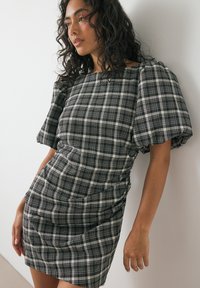 Ženska s kodrastimi temnimi lasmi, oblečena v obleko s pufastimi rokavi v črno-beli tartan vzorcu, se naslanja na belo steno.