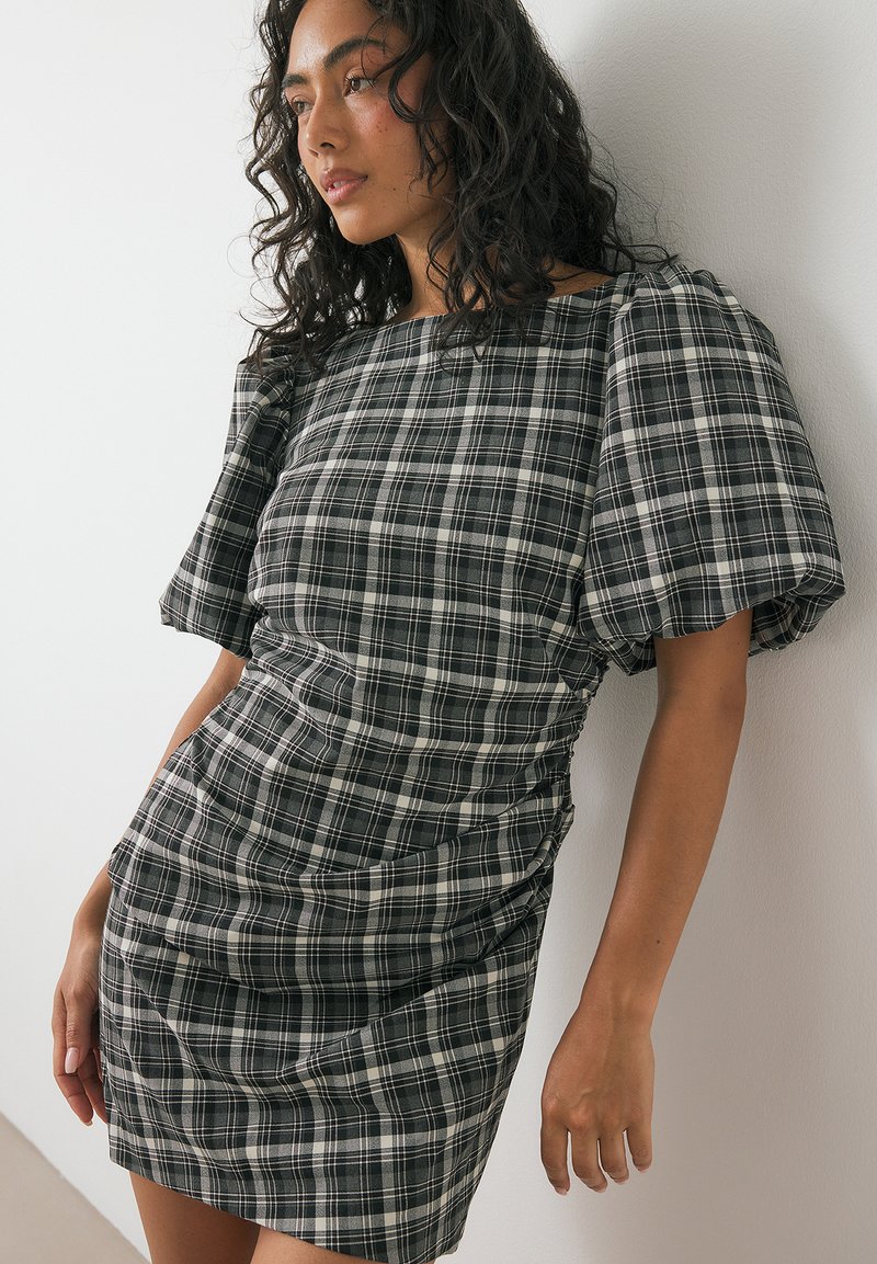 Ženska s kodrastimi temnimi lasmi, oblečena v obleko s pufastimi rokavi v črno-beli tartan vzorcu, se naslanja na belo steno.
