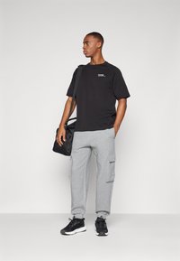 Młody mężczyzna stoi, ubrany w czarną koszulkę, szare spodnie dresowe typu cargo, czarne trampki, nosi czarną torbę na ramię na jednolitym tle.