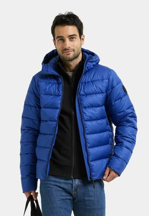 Man draagt een blauwe donsjas over een zwarte sweater met rits en blauwe spijkerbroek, houdt met zijn linkerhand een zwarte tasriem vast.