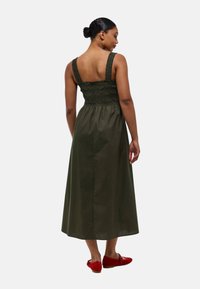 Robe maxi vert olive avec un corsage froncé et de larges bretelles, dotée d'une jupe fluide et accompagnée de chaussures rouges à enfiler.