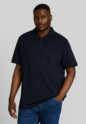 Young man with short hair wearing a dark navy blue polo shirt and blue jeans, standing with one hand in his pocket against a gray background.