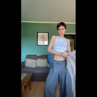 Un top cropped fără mâneci, de culoare albastră deschisă, asortat cu pantaloni largi gri. Fundalul include un perete verde și o canapea maronie.