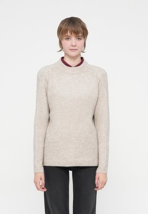 Beige ribbed sweater with a high neckline, long sleeves, and a slightly loose fit. Worn over a dark shirt, featured against a plain background.
