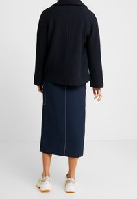 Zwarte wolmix jack met brede kraag, gecombineerd met een marineblauwe rok tot op de knie met contrasterende witte stiksel. Gekozen voor beige sneakers.