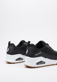 Zapatillas deportivas negras con parte superior de malla, entresuelo blanco y cojín de aire translúcido. Presenta una suela de goma marrón claro y el logotipo de Skechers.