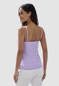 Lilafarbener Tank-Top mit dünnen Trägern, versehen mit einem strukturierten Stoffdesign. Angezeigt an einem Model, kombiniert mit weißen Hosen, von hinten betrachtet.