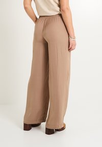 Pantalones de pierna ancha beige con una tela ligera y texturizada y una cintura elástica. Terminados con un dobladillo limpio y combinados con zapatos de tacón marrones.