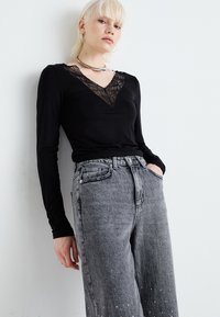 Top negro de manga larga con detalle de encaje en el escote en V, acompañado de jeans grises de cintura alta con textura desgastada y adornos en el dobladillo.