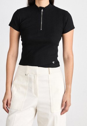 Žena má na sobě černý žebrovaný top s krátkými rukávy a předním zipem a bílé kalhoty s vysokým pasem a velkými kapsami.