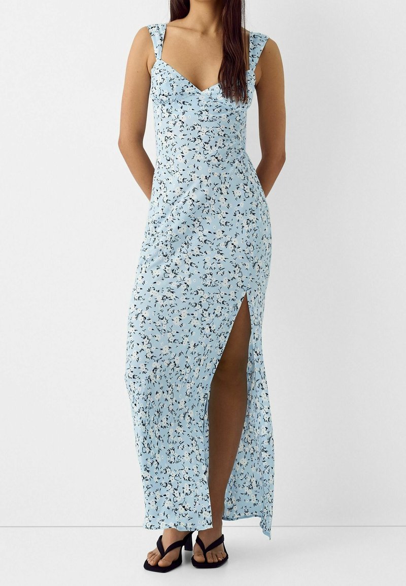 Femme portant une robe longue fleurie bleu clair avec une fente haute sur la cuisse et des sandales à talons noires, debout devant un fond blanc uni.