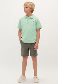 Polo verde menta a maniche corte, con colletto e decorazione ricamata. Indossato con pantaloni corti verde oliva e sneakers chiare.