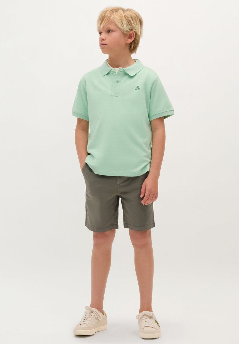 Polo verde menta a maniche corte, con colletto e decorazione ricamata. Indossato con pantaloni corti verde oliva e sneakers chiare.