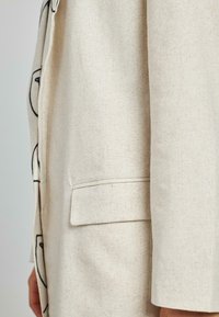 Cappotto in lino beige con dettagli a contorno neri, chiusura a bottone singolo e tasca laterale, caratterizzato da un taglio pulito e dritto e da un tessuto testurizzato.