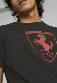 Czarna bawełniana koszulka z dużym czerwonym logo Ferrari, przedstawiającym galopującego konia w środku tarczy oraz napisem "S F" na dole.